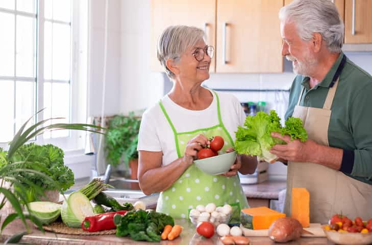 An elderly couple in their kitchen surrounded by fruits and vegetables to eat healthier.