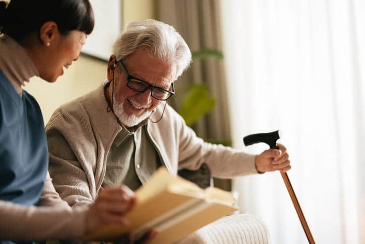 An Elderly Man Reading A Book With A Nurse At A Nursing Home.