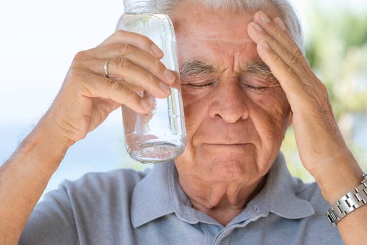 A Man Holding His Head During A Heat-Related Illness. In One Hand Is A Glass Of Water Pressed To His Temple.