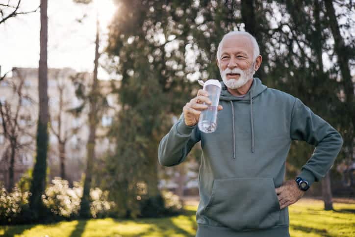 A Man Wearing A Hoodie While Smiling And Drinking Water Outside As He Enjoys The Weather.